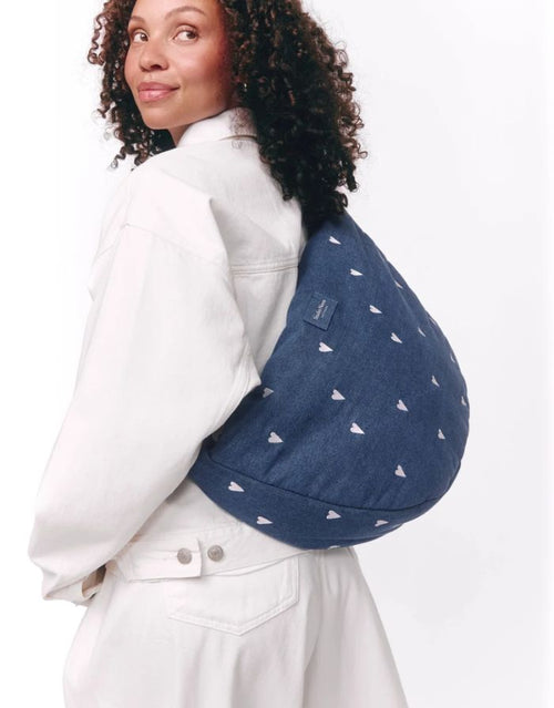 Studio Noos Cross Body Umhängetasche aus dunkelblauem gepolstertem Denim mit gestickten weißen Herzen und Platz für einen 13-Zoll-Laptop getragen.