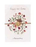 Schmuck-Postkarte New Home von A Beautiful Story mit „happy new home“-Hausmotiv und Armband mit vergoldetem Herzanhänger.