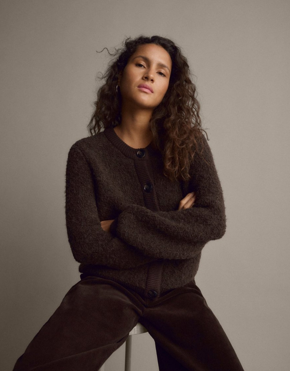 Dunkelbraune Taile Nerida Strick Jacke von MSCH Copenhagen aus weichem Woll- und Alpakamix mit Rundhalsausschnitt, dunklen Knöpfen und gerippten Abschlüssen am Model auf einem Stuhl