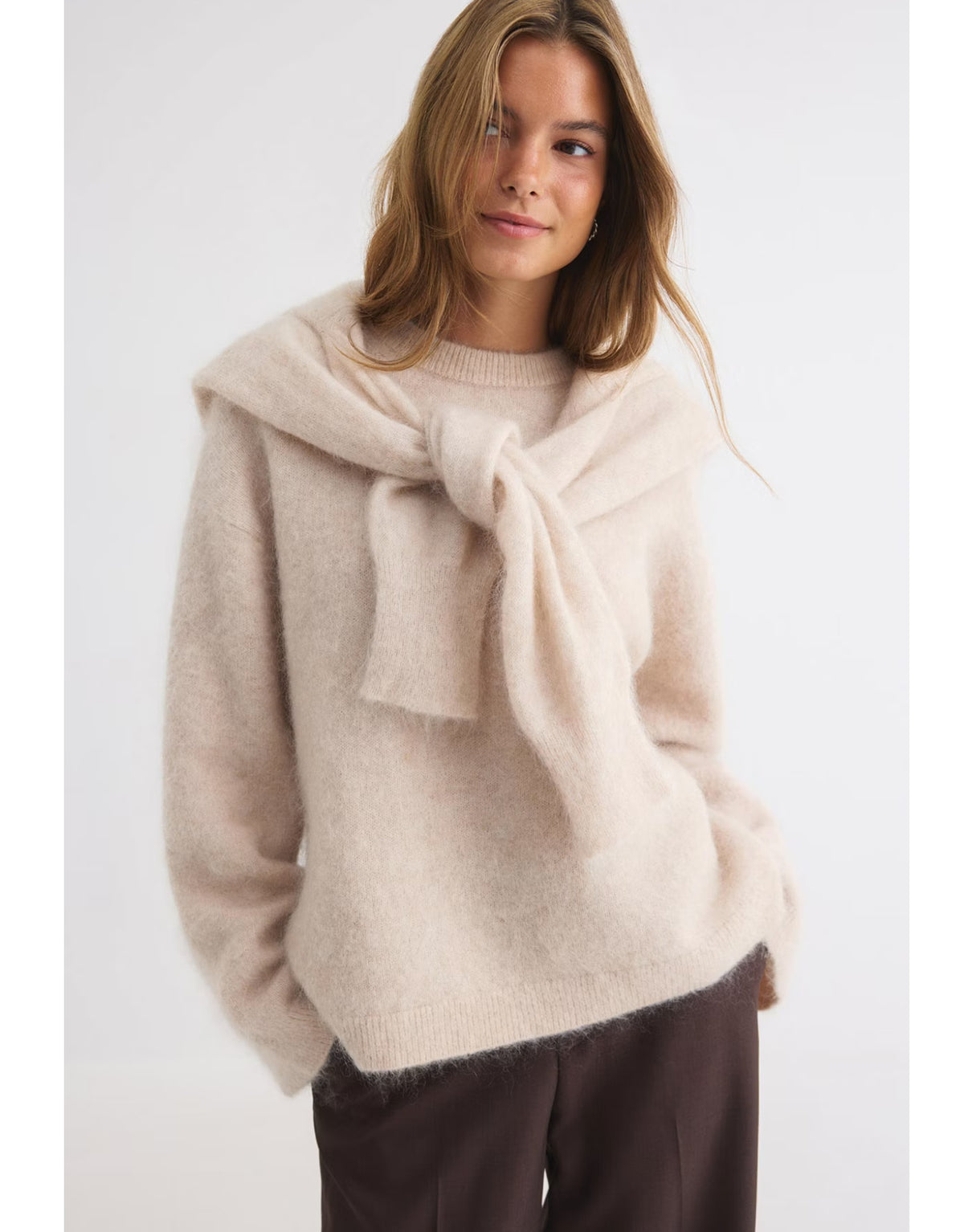 Beiger Mohair Pullover von Gina Tricot mit feiner, flauschiger Strickstruktur, lockerer Passform, langen Ärmeln und klassischem Rundhalsausschnitt am Model von vorne mit Pullover um die Schultern