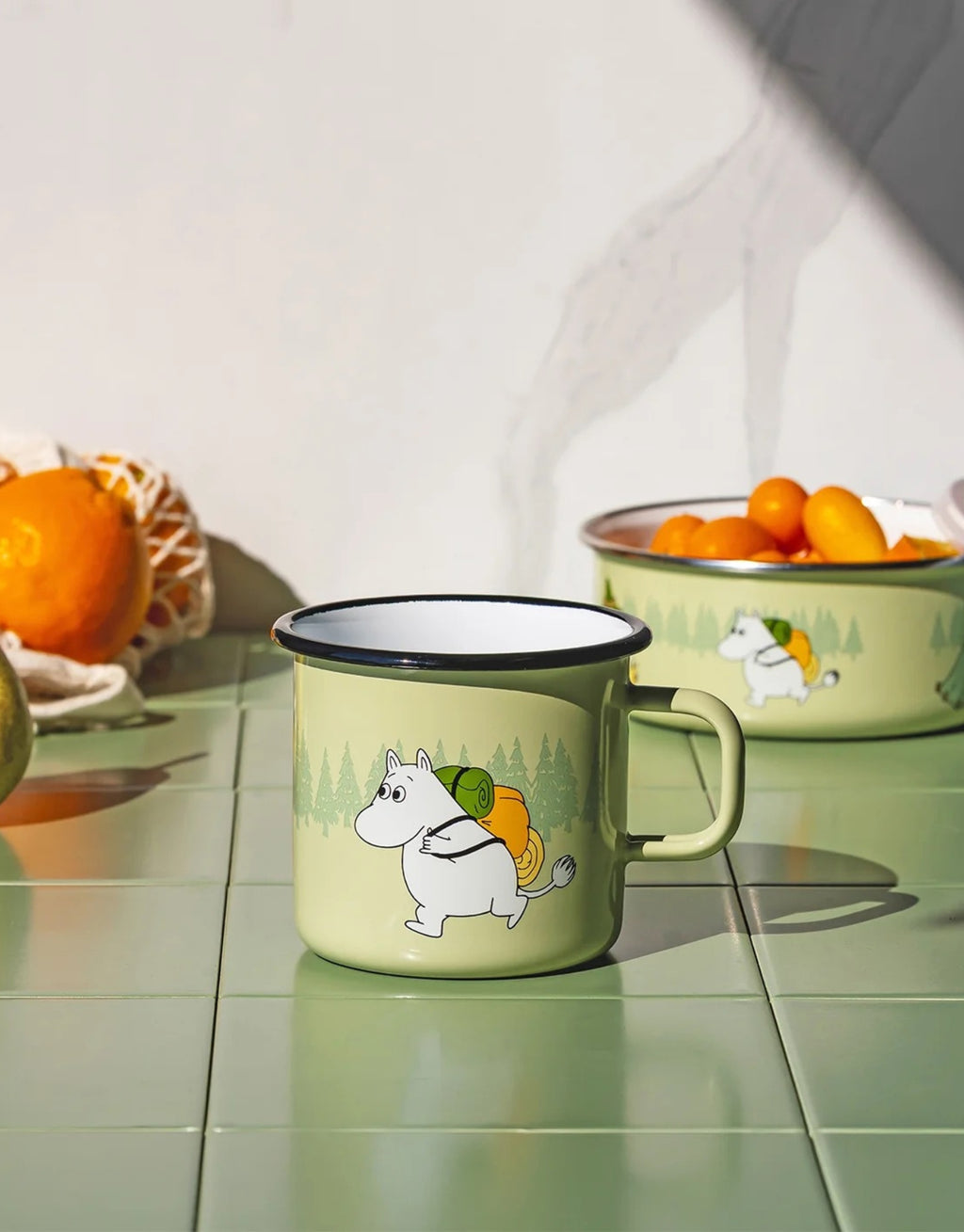 Moomin Emaille-Becher Campers von Muurla in Grün, dekorativer Trinkbecher aus emailliertem Stahl mit Henkel und liebevollem Moomin-Ausflugsmotiv, 550 ml Fassungsvermögenauf auf grünen Fliesen