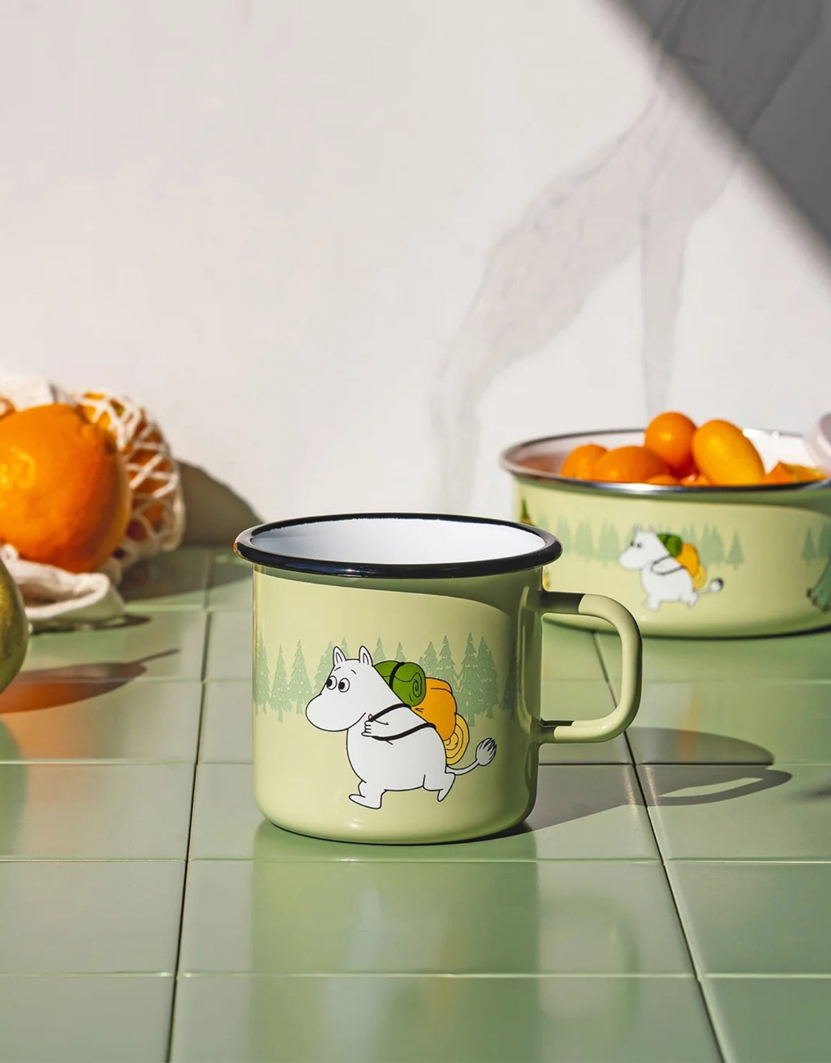 Moomin Emaille-Becher Campers von Muurla in Grün, dekorativer Trinkbecher aus emailliertem Stahl mit Henkel und liebevollem Moomin-Ausflugsmotiv, 550 ml Fassungsvermögenauf auf grünen Fliesen