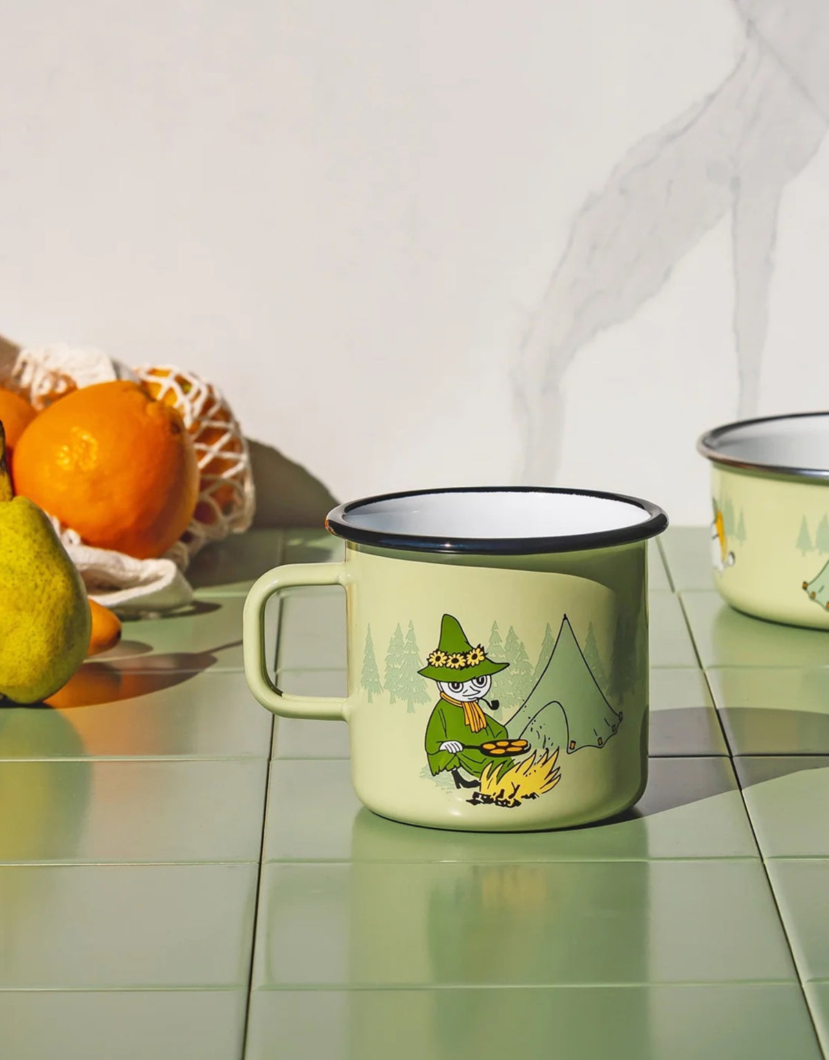 Moomin Emaille-Becher Campers von Muurla in Grün, dekorativer Trinkbecher aus emailliertem Stahl mit Henkel und liebevollem Moomin-Ausflugsmotiv, 550 ml Fassungsvermögen auf Fliesen vo der anderen Seite