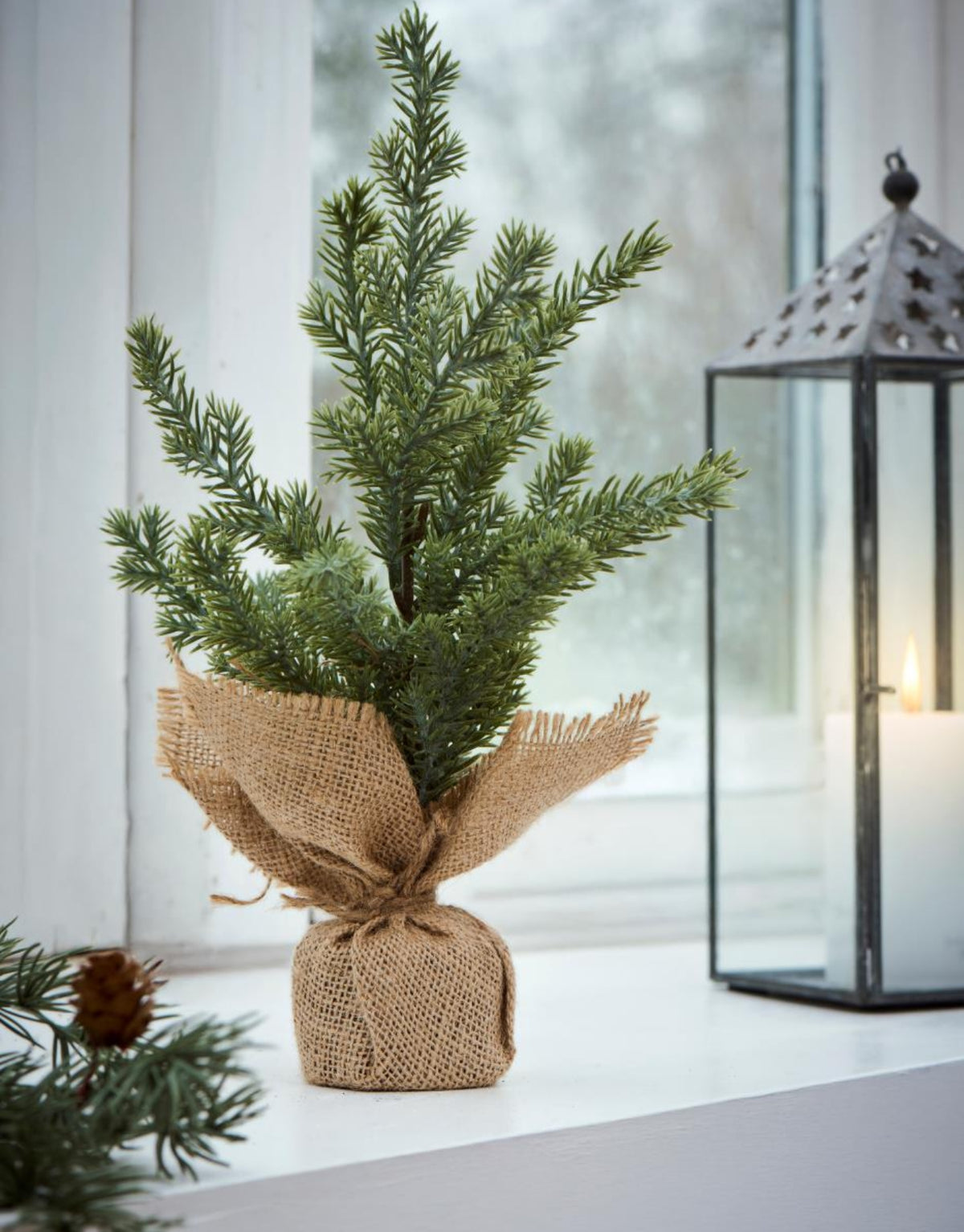 Kleine grüne Kunsttanne Cedar von Stillenat by Ib Laursen mit naturgetreuen Zweigen und Jutefuß, 32 cm hoch auf einer Fensterbank.