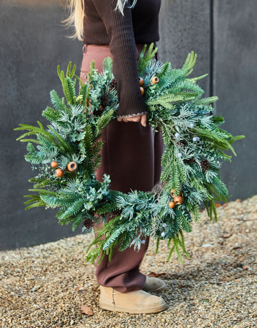 Weihnachtskranz Deco von House Doctor in Grün, 68 cm großer künstlicher Weihnachtskranz mit üppigen tiefgrünen Zweigen, dekorativen Fichtenzapfen und stabilem Eisenrahmen in der Hand einer Frau