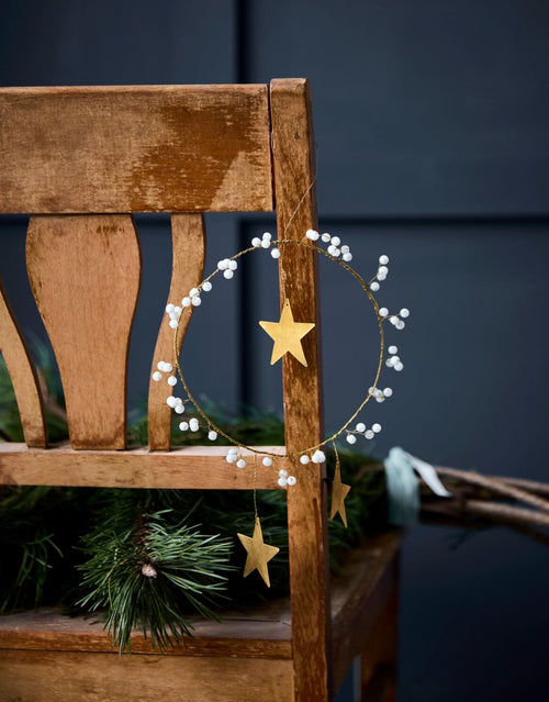  Handgefertigtes Weihnachtsornament von House Doctor als runder Kranz aus Messing mit weißen Glasperlen und drei Sternen an einem Stuhl
