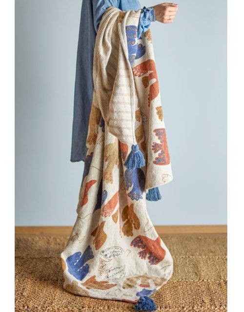 Anniversary Decke von Bloomingville in Beige mit botanischen und floralen Motiven in Blau und Braun sowie großen blauen Quasten, 160 × 130 cm über dem Arm eines Models