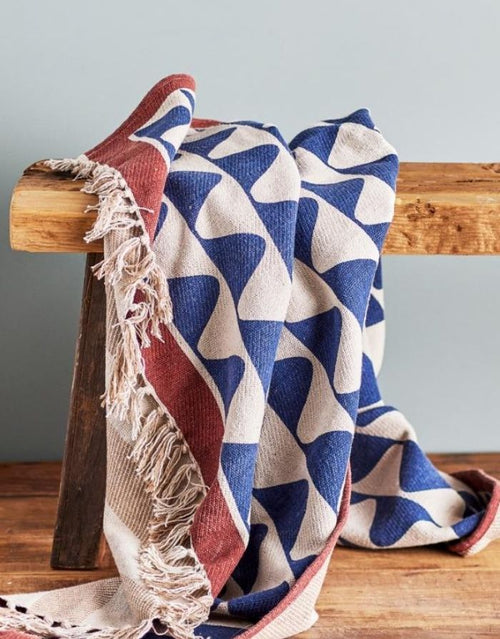 Bunte Decke Rimme von Creative Collection by Bloomingville mit blauem Wellenmuster, roten und beigen Akzenten und Fransen, 160 × 130 cm, auf einer Bank.